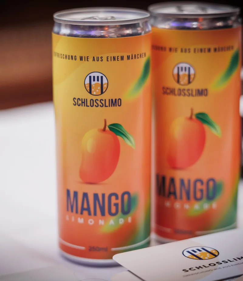 Zwei Dosen Schlosslimo Mango nebeneinander, dahinter ein Flyer mit dem Logo.